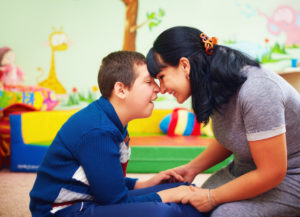 A child with cerebral palsy and their mother laugh, touching foreheads and holding hands in a colorful playroom.