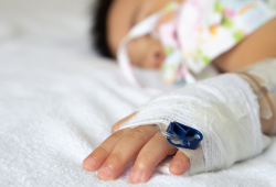 A young child lies flat in a hospital bed, head turned to the side, with a bandaged hand and an IV port.
