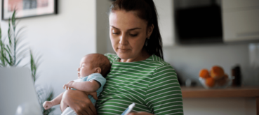 A mother holds a baby while reviewing information and taking notes at home.
