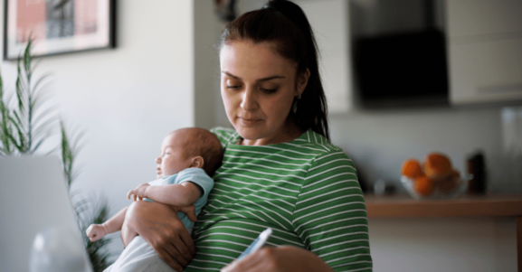 A mother holds a baby while reviewing information and taking notes at home.