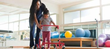 A physical therapist supports a child with cerebral palsy during strengthening exercises for CP child, using a walker in a well-equipped therapy room.