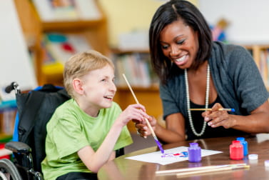 A special education teacher and child who uses a wheelchair laugh and paint together.