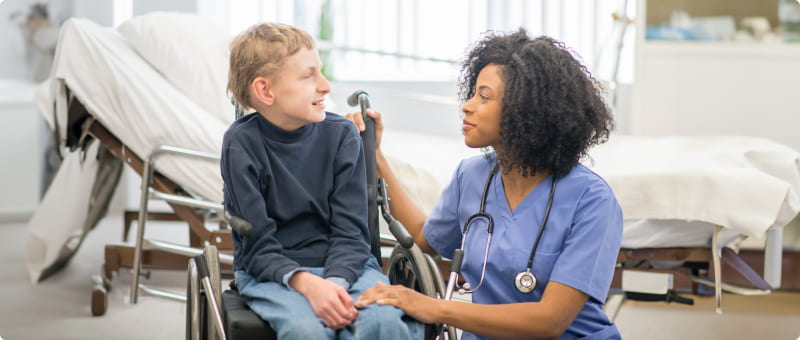 A child in a wheelchair and a health care provider talk and laugh in a hospital room.
