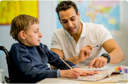 A child with cerebral palsy in a wheelchair works on a school assignment with an adult teacher assisting.
