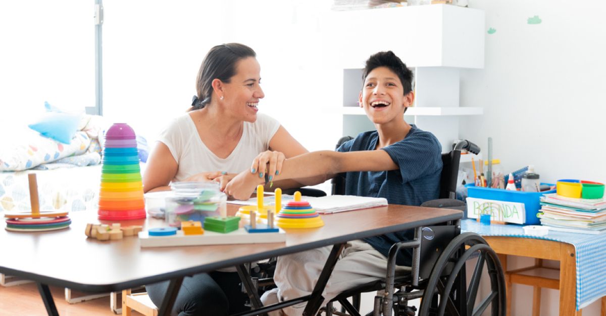 A woman who may be the child's mother or teacher, sits in a classroom, working with a wheelchair-bound child who presumably has cerebral palsy. Both are smiling.