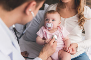 A doctor using a stethoscope to examine a baby with a pacifier, while the mother holds the baby's hand.