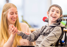 A smiling child with cerebral palsy in a wheelchair doing therapy exercises with a caregiver.