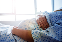A pregnant woman wearing a hospital gown lies in bed, gently holding her belly as soft daylight filters through a nearby window.