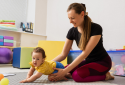 A caregiver assists a baby with crawling exercises on a padded floor in a brightly lit playroom.