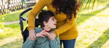 A mother leans down to hug her smiling young son, who is sitting in a wheelchair, while spending time together outdoors on a sunny day