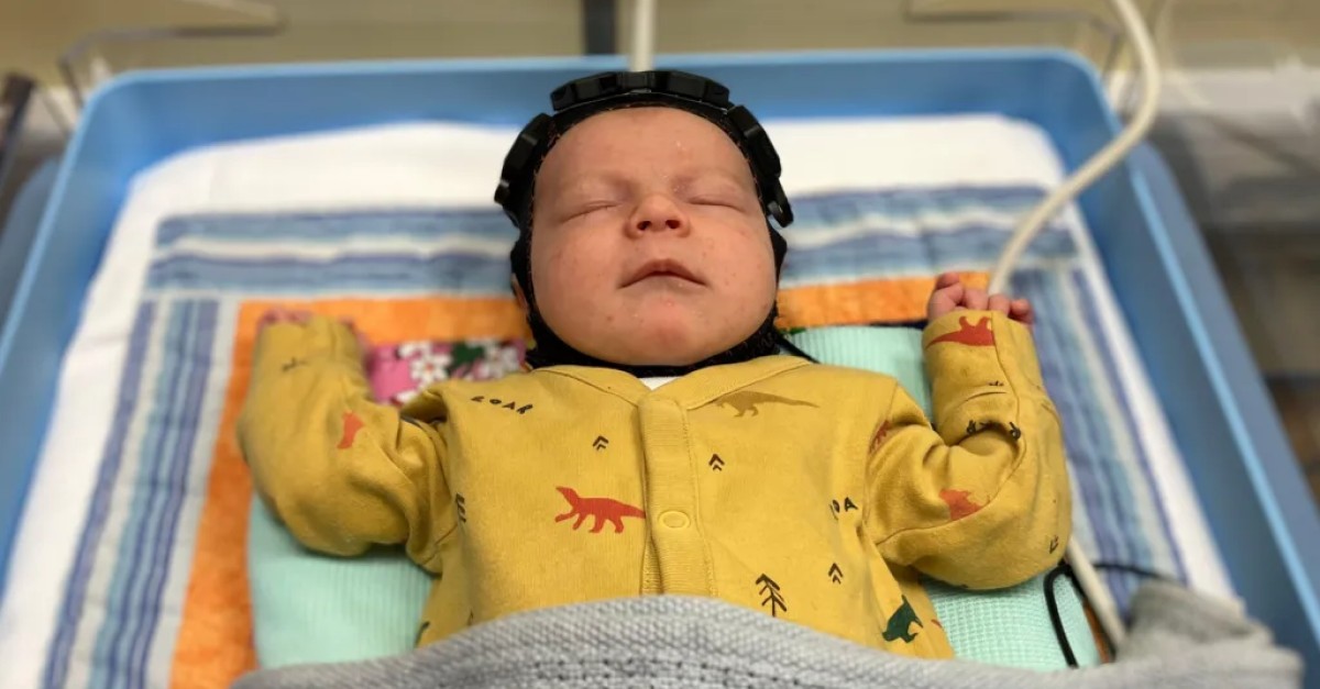 An infant rests in a cot and wears a cap designed to help with an early diagnosis of cerebral palsy and other birth injuries