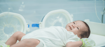 Newborn baby lying on a hospital bassinet, wearing a white onesie and monitoring bands, with medical equipment visible in the background