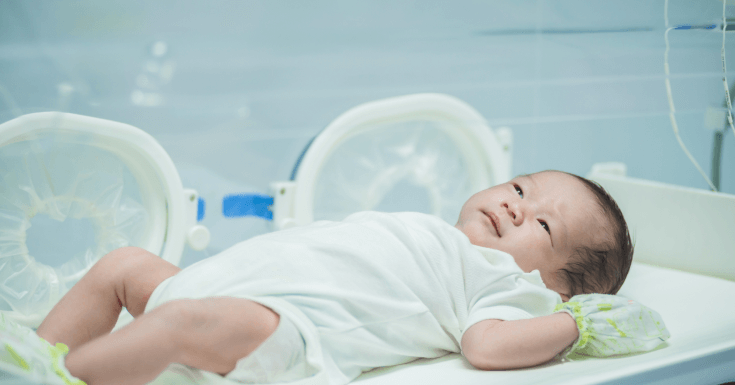 Newborn baby lying on a hospital bassinet, wearing a white onesie and monitoring bands, with medical equipment visible in the background
