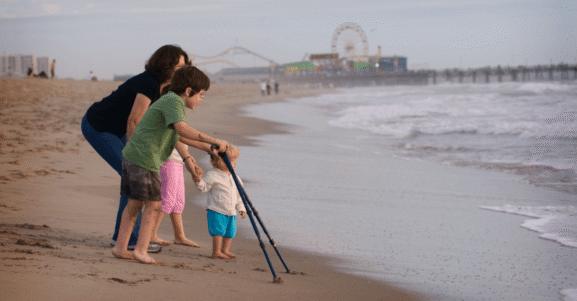 A family walks along the beach with a young child using forearm crutches.