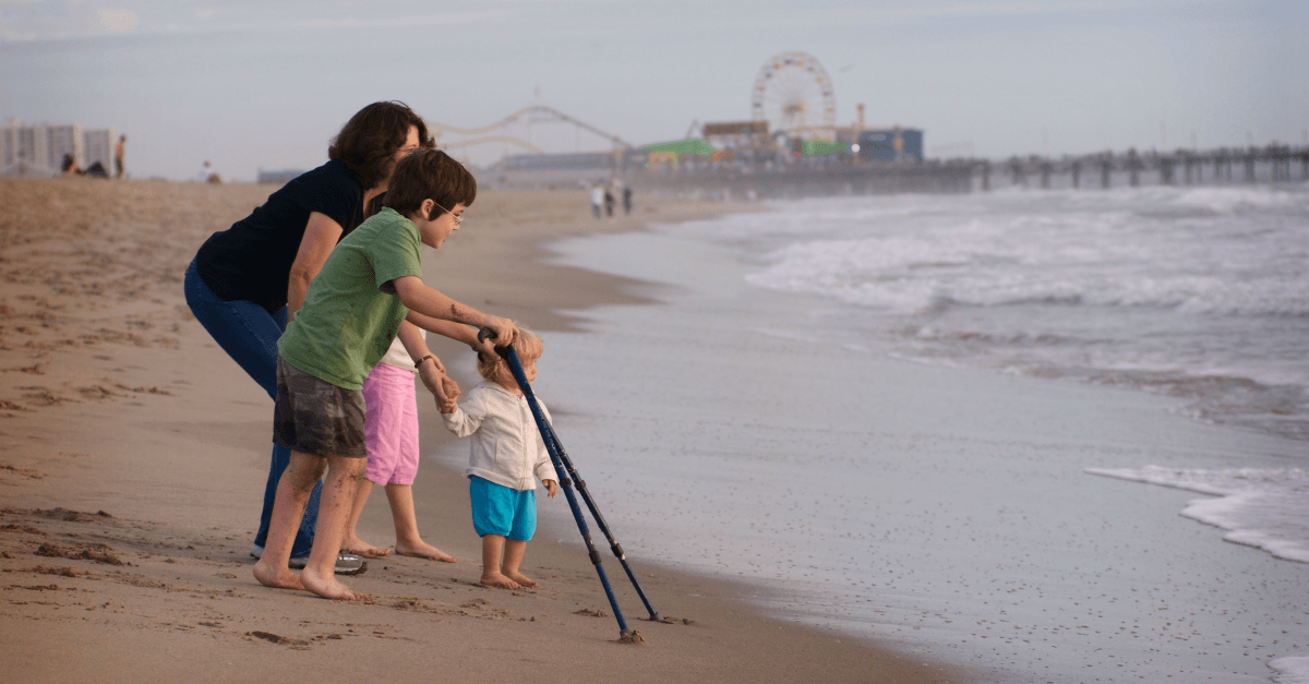 A family walks along the beach with a young child using forearm crutches.