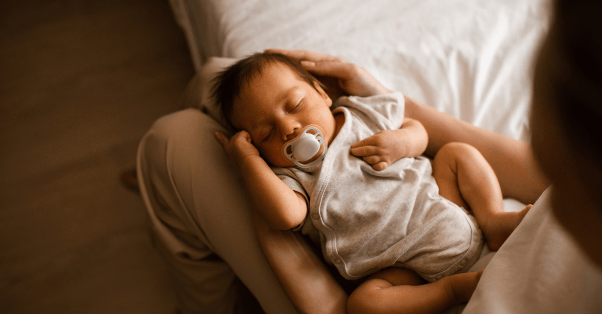 A sleeping baby with a pacifier rests in their mother's lap.