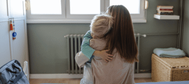 A parent sits on a bed holding and hugging a young child.