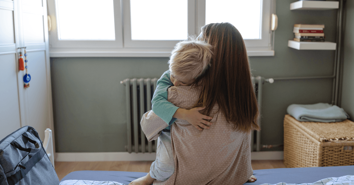 A parent sits on a bed holding and hugging a young child.