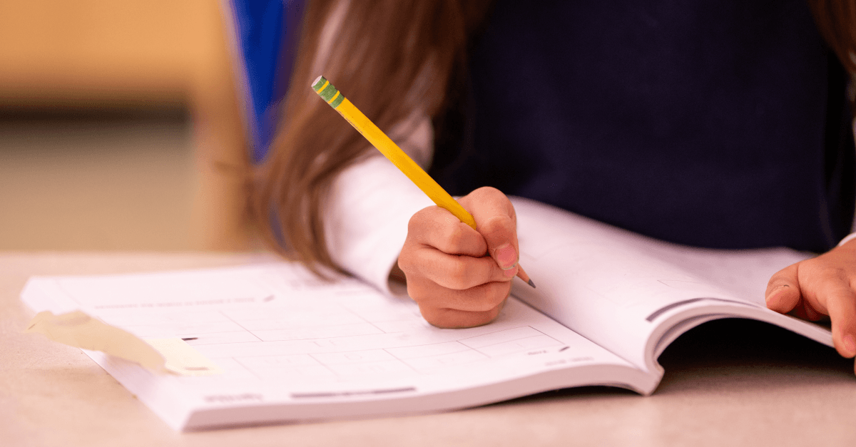 A student writes with a pencil in an open workbook.