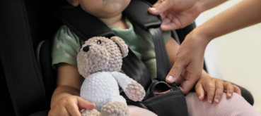 An adult adjusts a child's car seat harness while the child holds a stuffed animal.