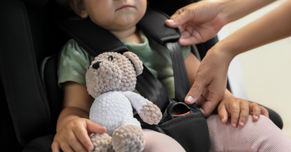 An adult adjusts a child's car seat harness while the child holds a stuffed animal.