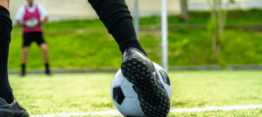 A soccer player rests one foot on a ball while another player stands near the goal.